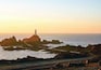 A lighthouse on a rocky outcrop with a road leading to it, surrounded by calm sea and clear sky.