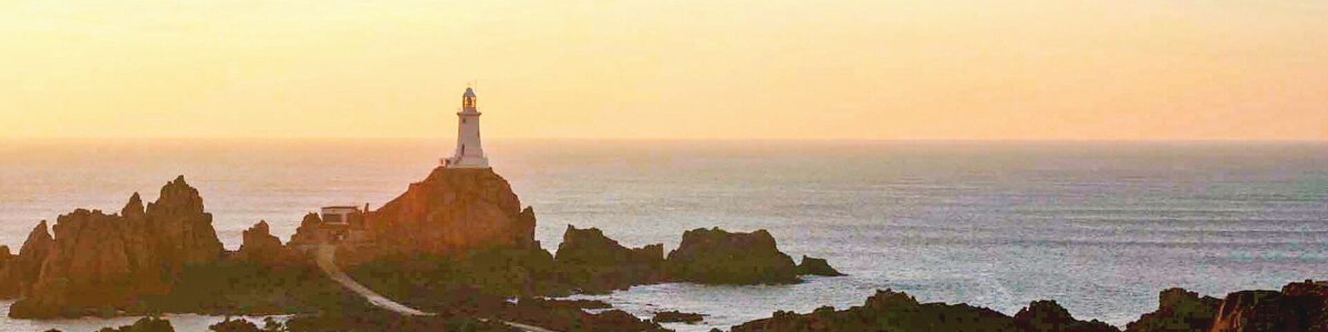 Un faro sobre un promontorio rocoso con una carretera que lo lleva, rodeado de mar tranquilo y cielo despejado.