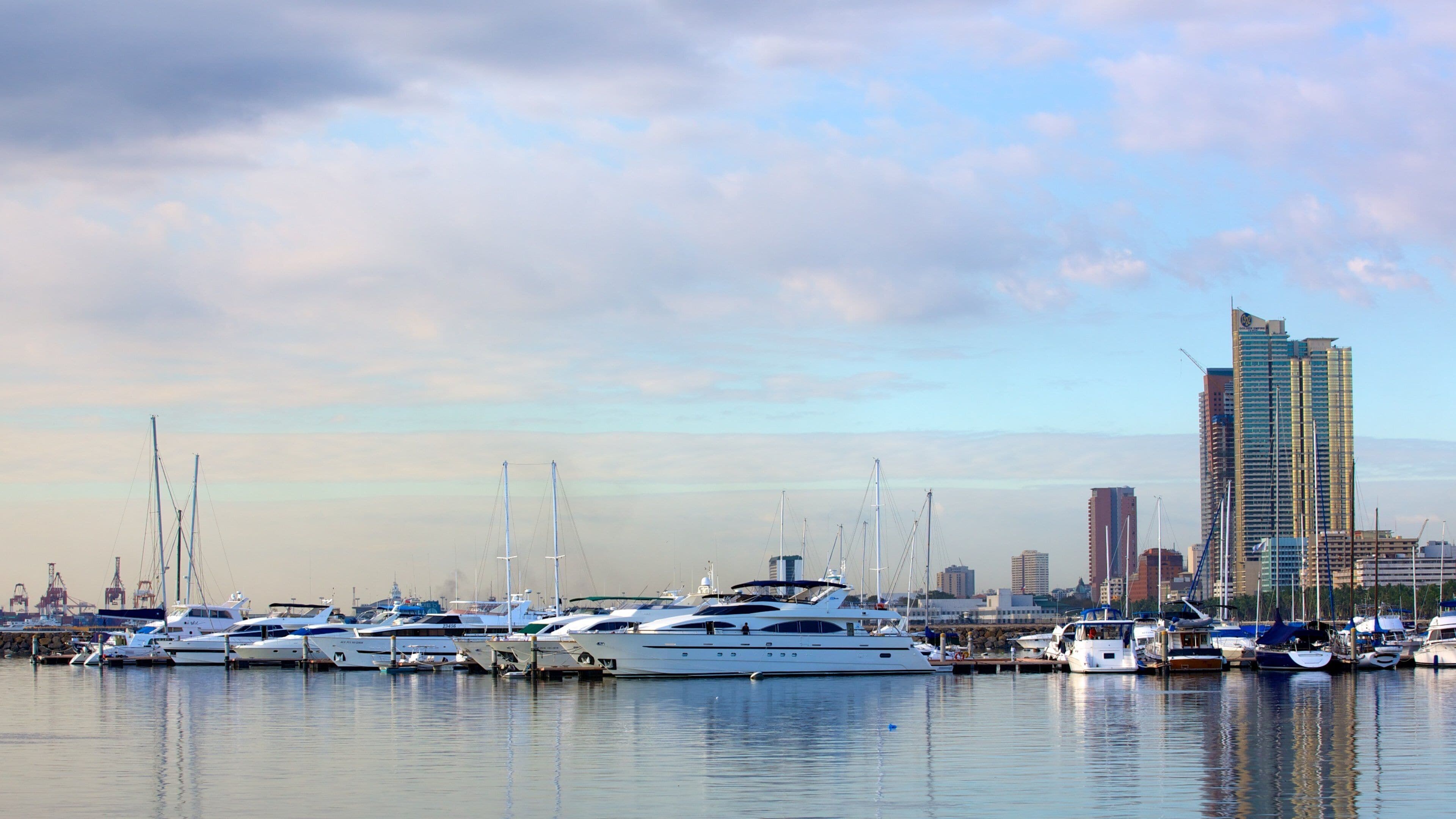 A marina with several yachts docked, a city skyline in the background, and a partly cloudy sky.