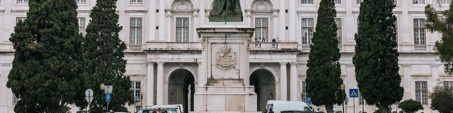 A statue in front of a large white building with columns and a balcony. There are tall trees on either side of the statue.