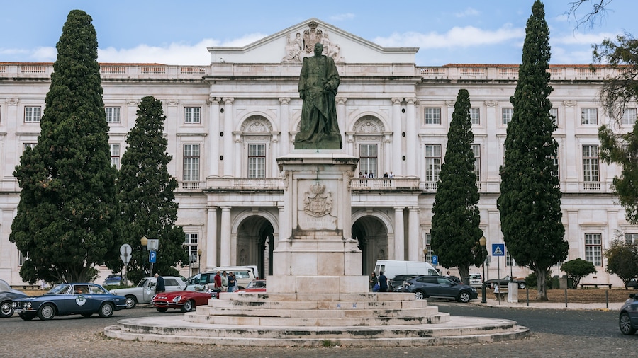 Una estatua frente a un gran edificio blanco con columnas y un balcón. Altas palmeras flanquean la estatua.