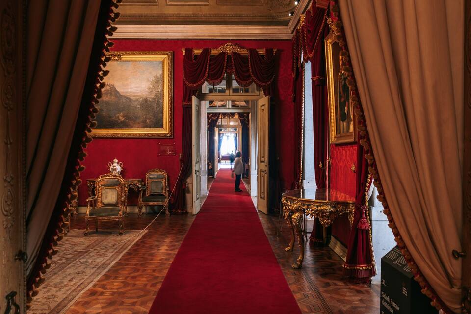 A grand hallway with a red carpet, ornate furniture, and a large painting on the wall.