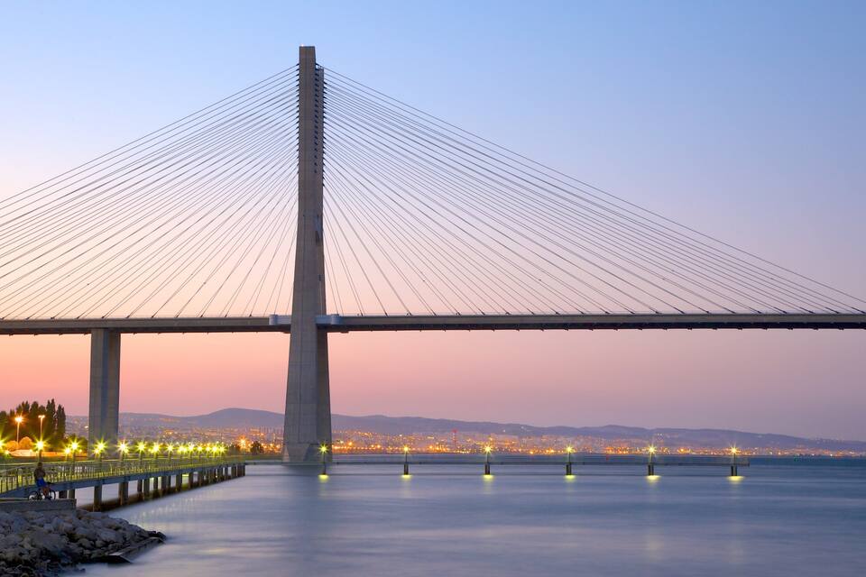 Eine Schrägseilbrücke in der Abenddämmerung mit Stadtlichtern im Hintergrund.