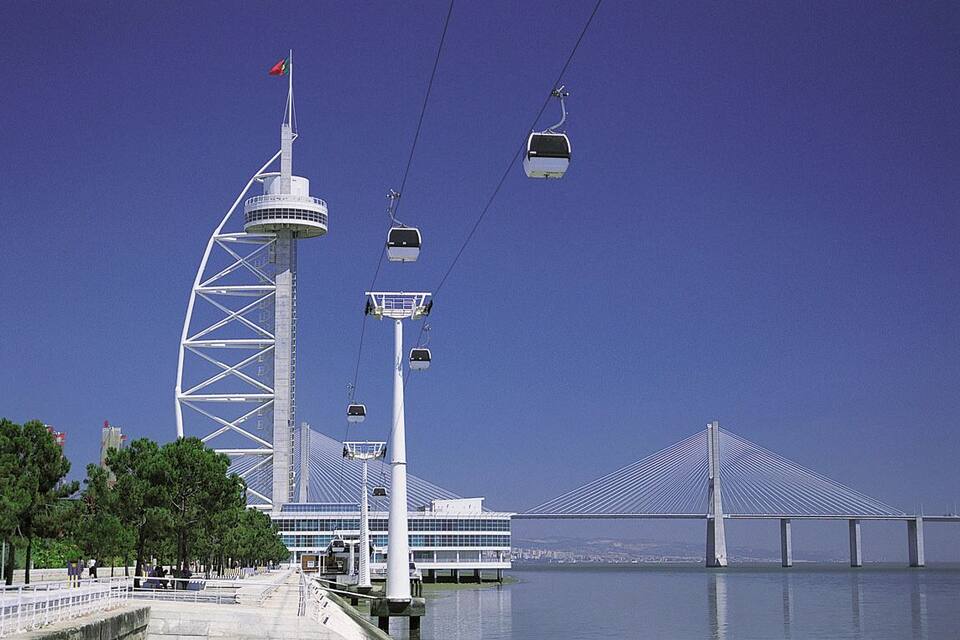 A modern tower with a flag, a cable car system, a bridge, and a waterfront.