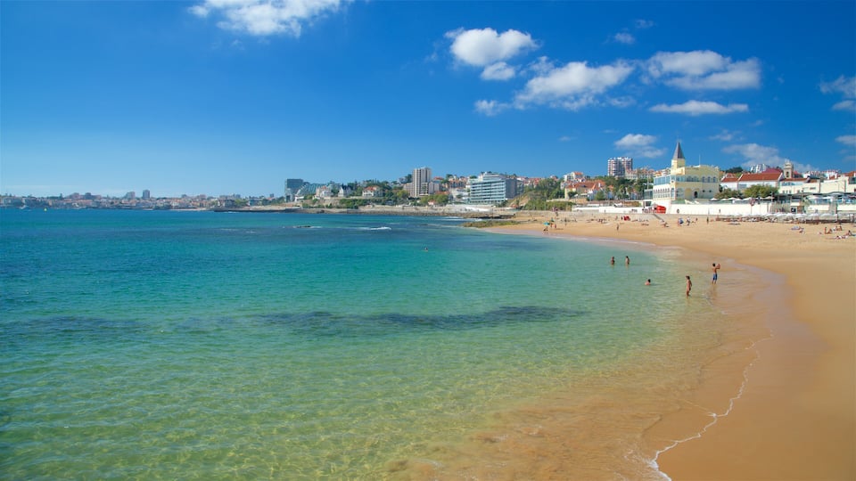 Una playa con agua turquesa cristalina, una orilla arenosa y un paisaje urbano con edificios y una iglesia al fondo.
