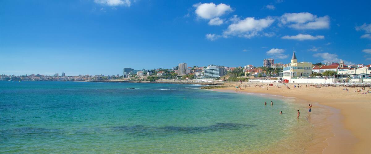 A beach with clear turquoise water, a sandy shore, and a cityscape with buildings and a church in the background.