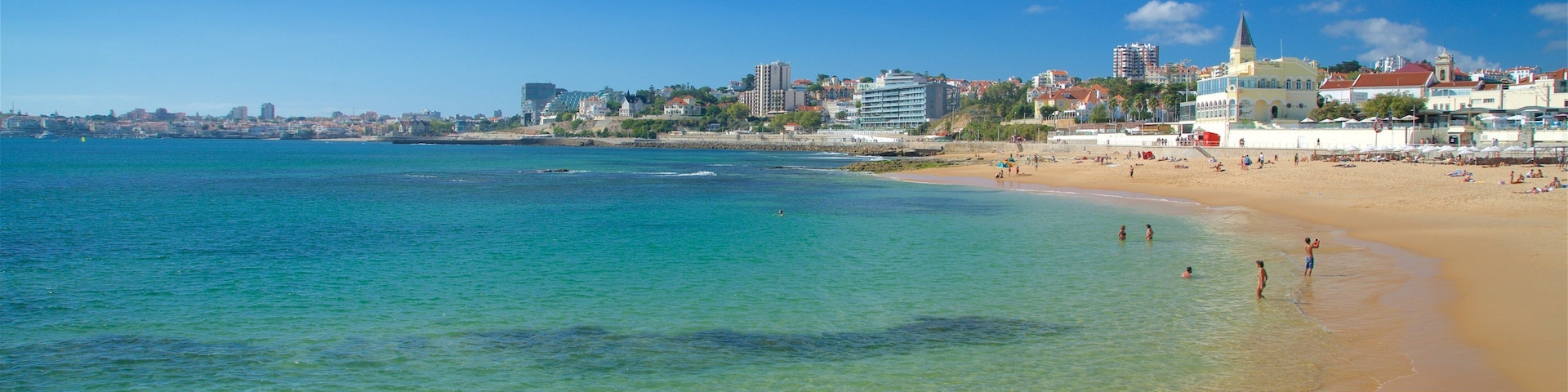 A beach with clear turquoise water, a sandy shore, and a cityscape with buildings and a church in the background.