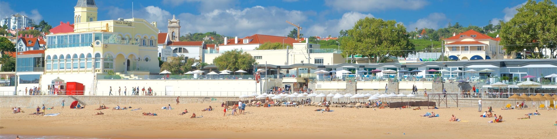 A beach with people sunbathing and swimming, a church with a bell tower, and a coastal town with buildings and umbrellas.