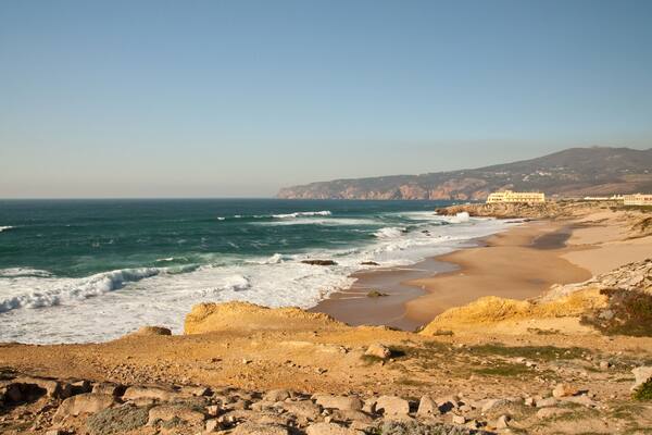 A coastal landscape with a sandy beach, rocky outcrops, and a building on the shore.