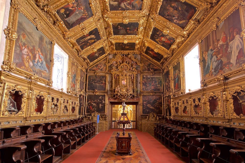 An ornate church interior with gold detailing, wooden pews, and religious paintings on the walls and ceiling.