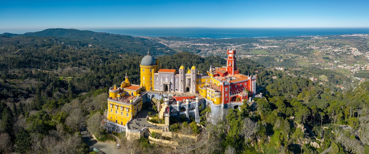 A castle with yellow and red buildings, surrounded by forest and overlooking a coastal town.