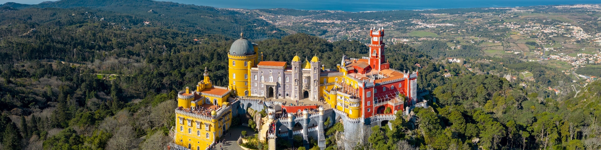 A castle with yellow and red buildings, surrounded by forest and overlooking a coastal town.