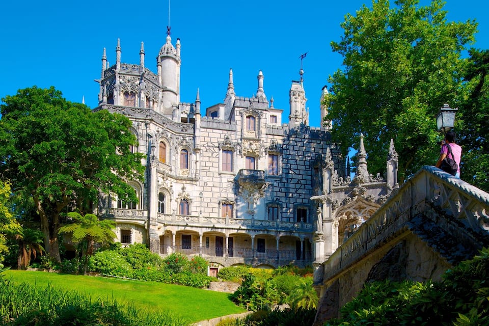 Un castillo histórico con arquitectura ornamentada, rodeado de exuberante vegetación y un puente de piedra.