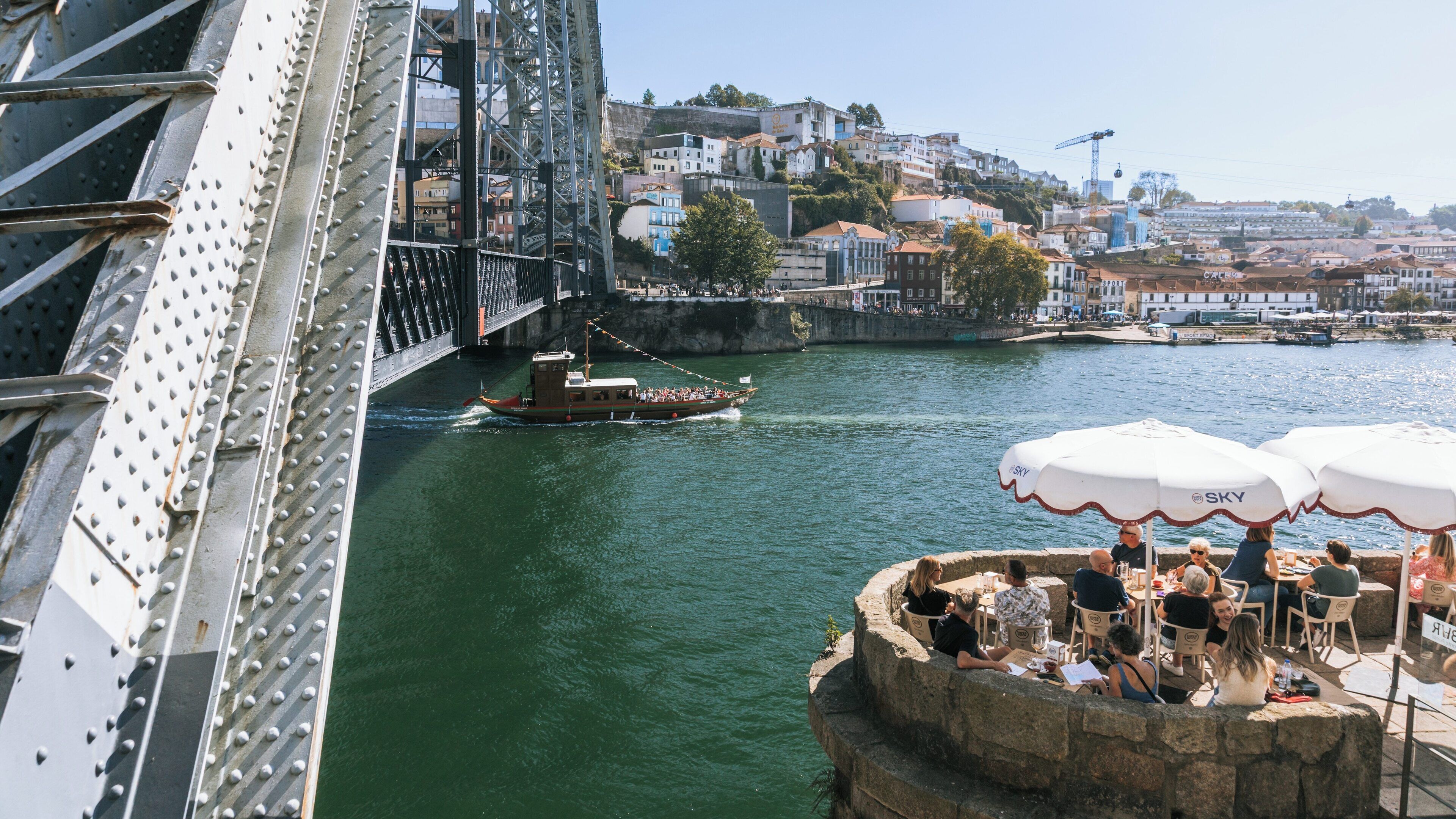 View of Dom Luis I Bridge in Sao Nicolau, Porto District, Portugal with waterfront dining and a boat cruising on the Douro River