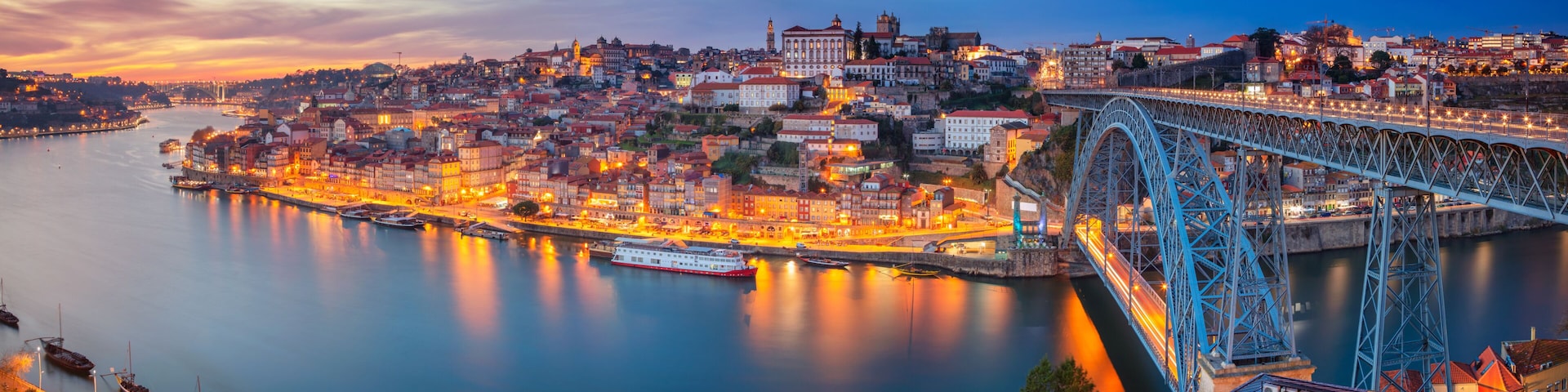 Une vue sur une ville au crĂ©puscule, avec un pont imposant enjambant une riviĂšre, des bĂątiments illuminĂ©s et une surface dâeau calme qui reflĂšte les lumiĂšres.