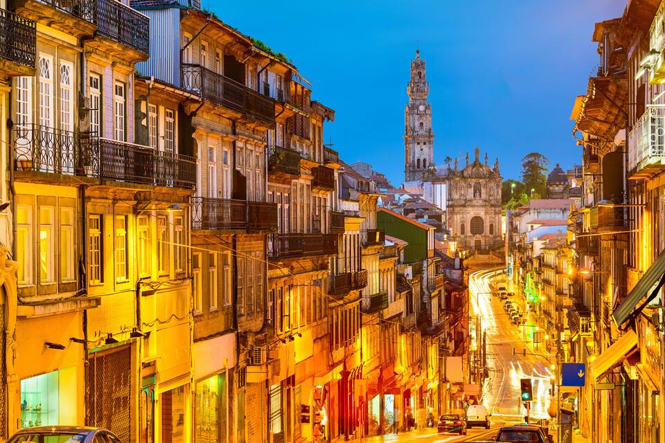 Uma rua histórica numa cidade europeia ao entardecer, com edifícios iluminados, uma torre do relógio imponente e uma catedral ao fundo.