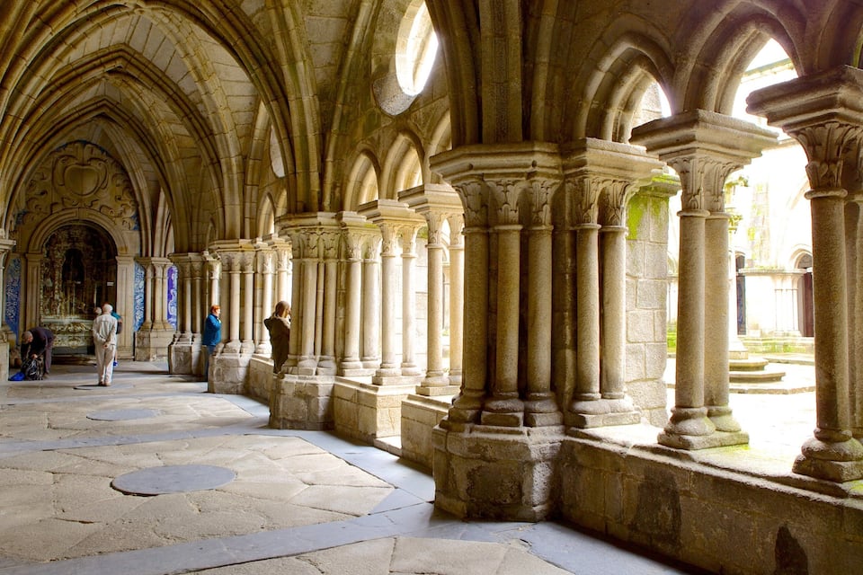 A Gothic-style cloister with stone columns and arches, a stone floor, and a view of the outside.
