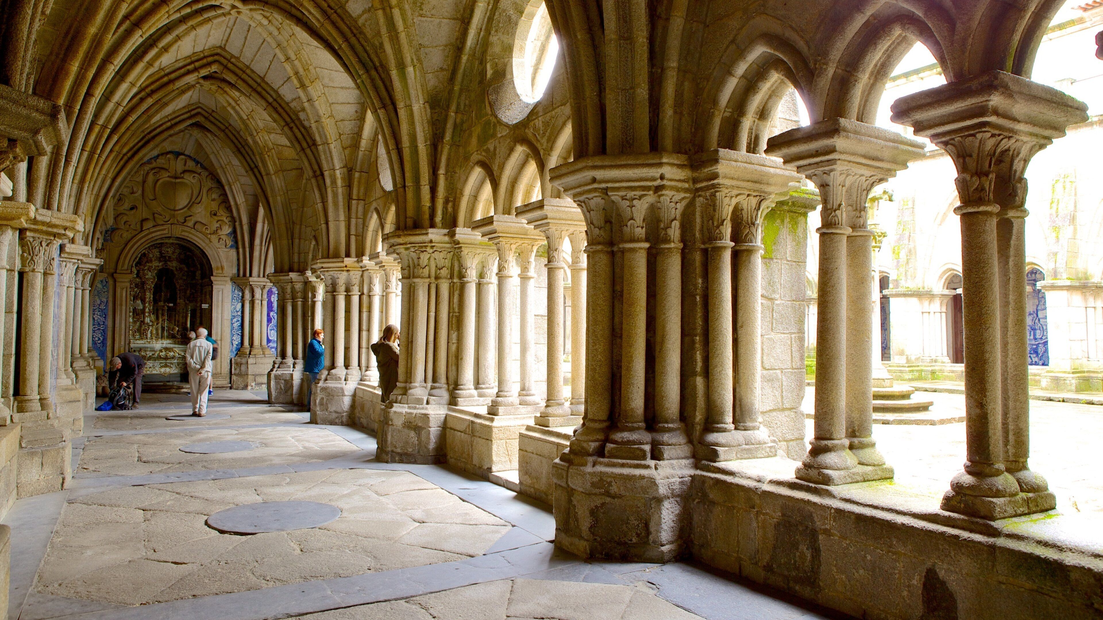 A Gothic-style cloister with stone columns and arches, a stone floor, and a view of the outside.