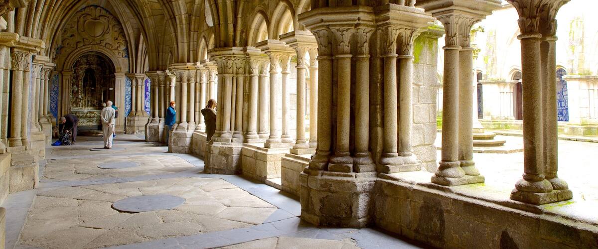 A Gothic-style cloister with stone columns and arches, a stone floor, and a view of the outside.