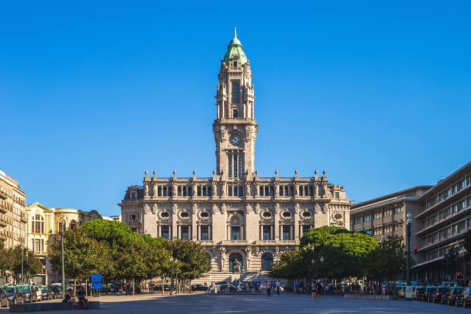 Um edifício histórico com uma torre do relógio, cercado por árvores e uma praça com pessoas.