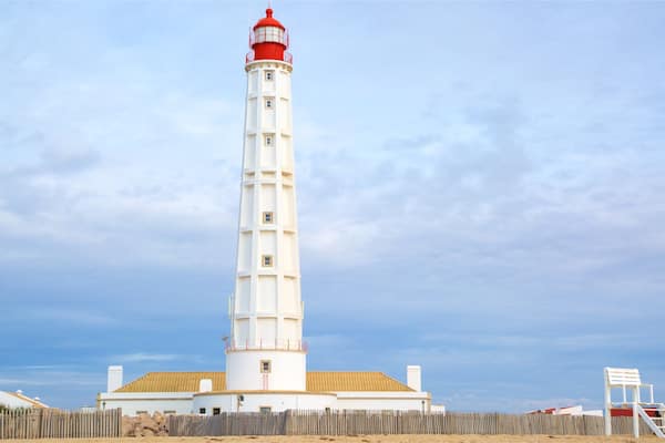 A tall white lighthouse with a red top, surrounded by a wooden fence and a white building with a yellow roof.