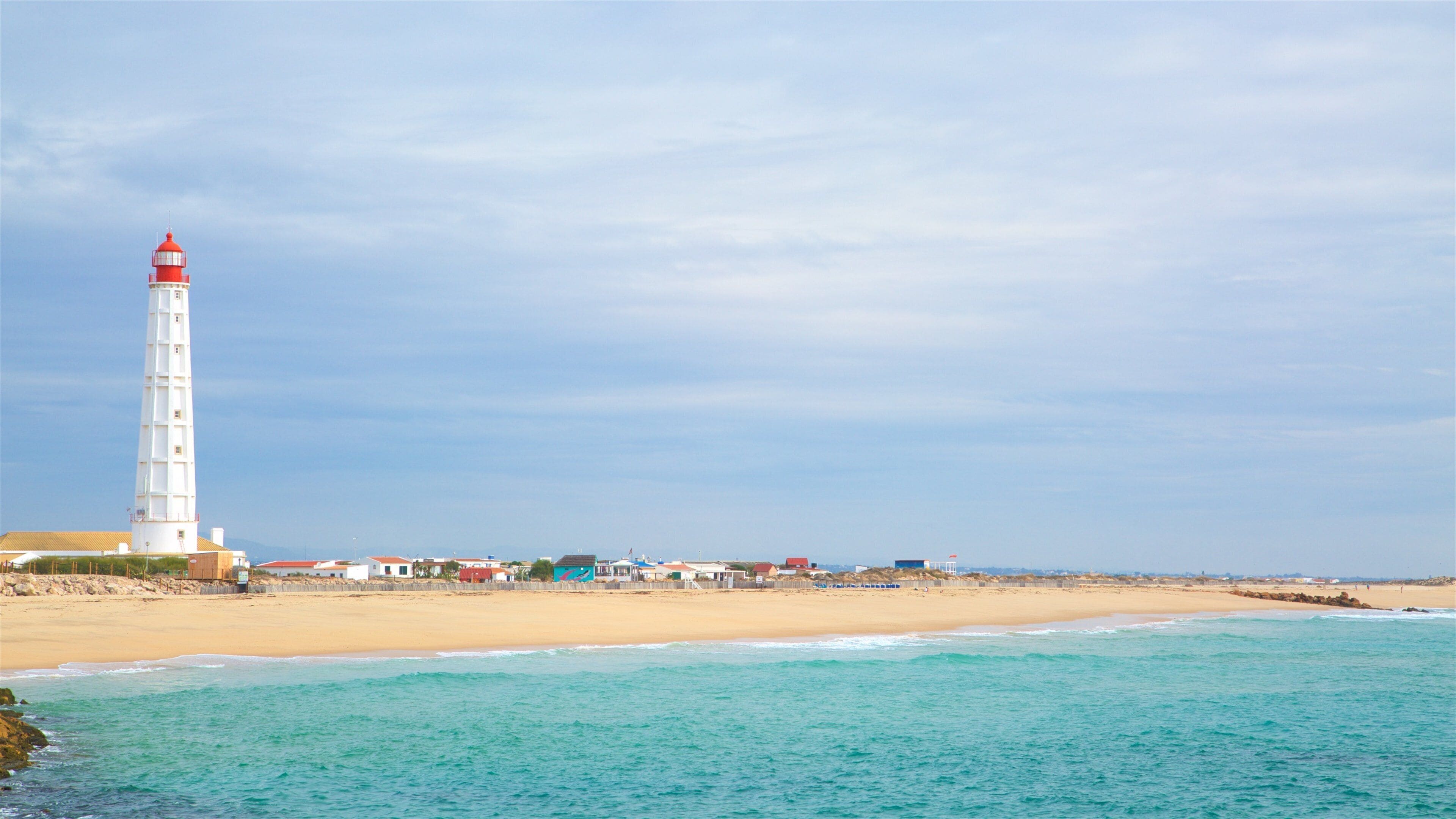A coastal scene with a white lighthouse, sandy beach, and turquoise waters.