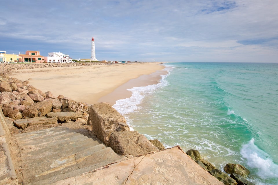 Un paysage côtier avec une plage de sable, une digue rocheuse et un phare visible au loin.