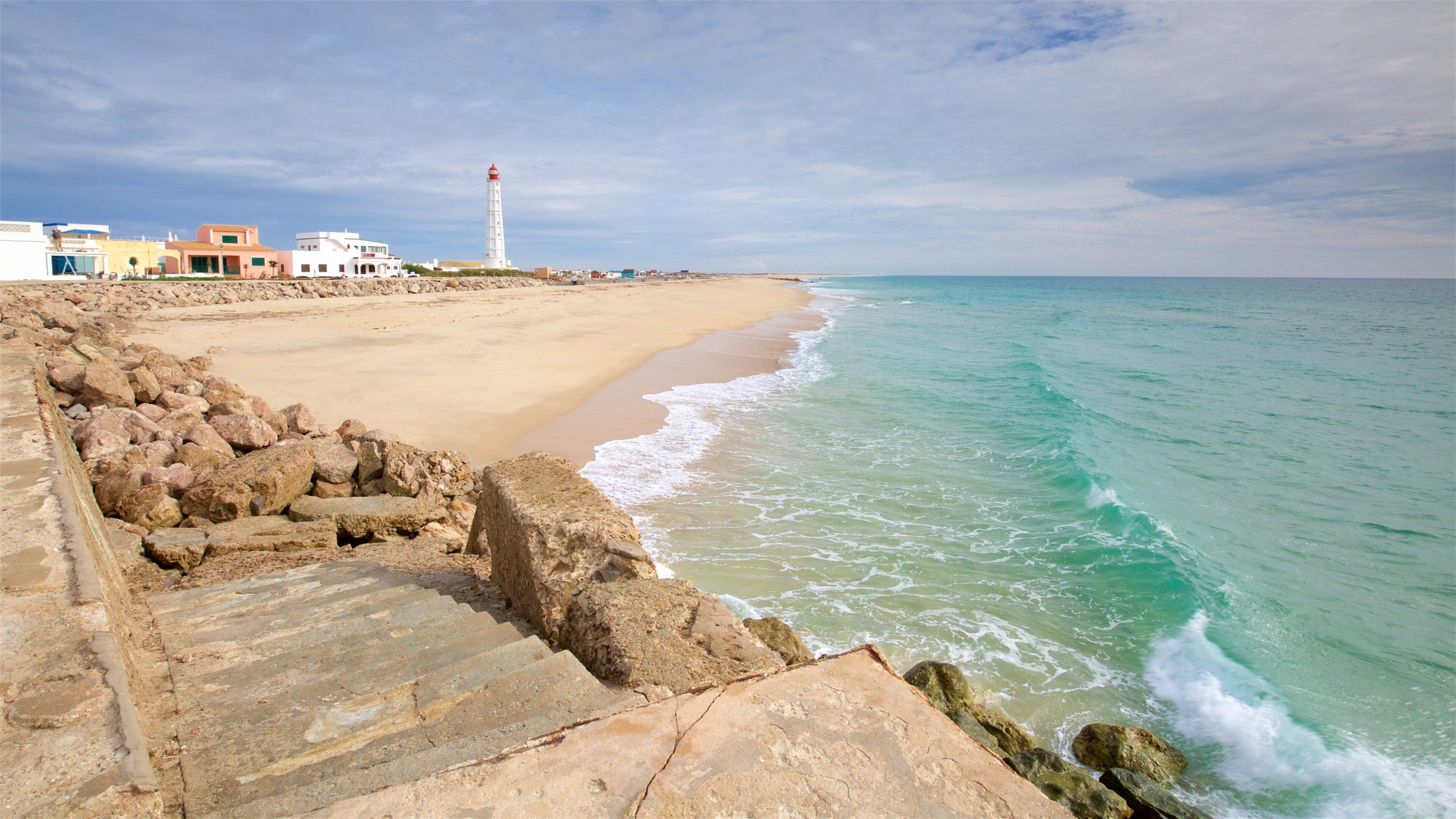 A coastal scene with a sandy beach, a rocky breakwater, and a lighthouse in the distance.
