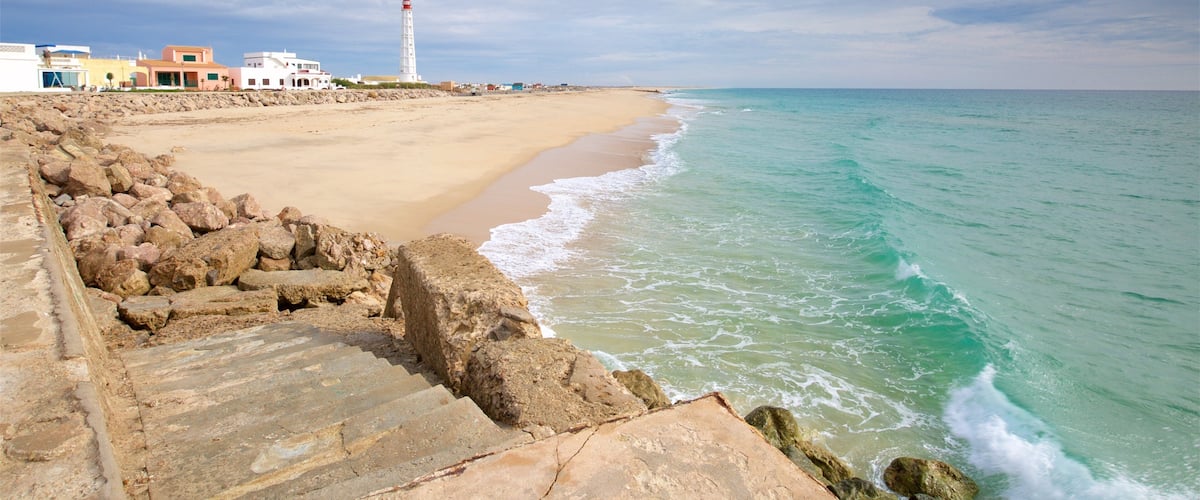 A coastal scene with a sandy beach, a rocky breakwater, and a lighthouse in the distance.