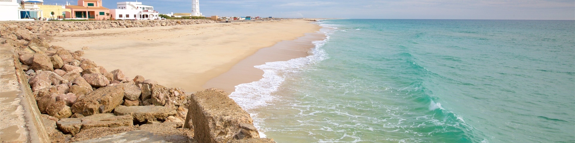 A coastal scene with a sandy beach, a rocky breakwater, and a lighthouse in the distance.