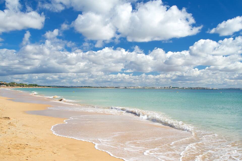 Uma praia com águas turquesa cristalinas, areal arenoso e silhueta urbana ao longe.