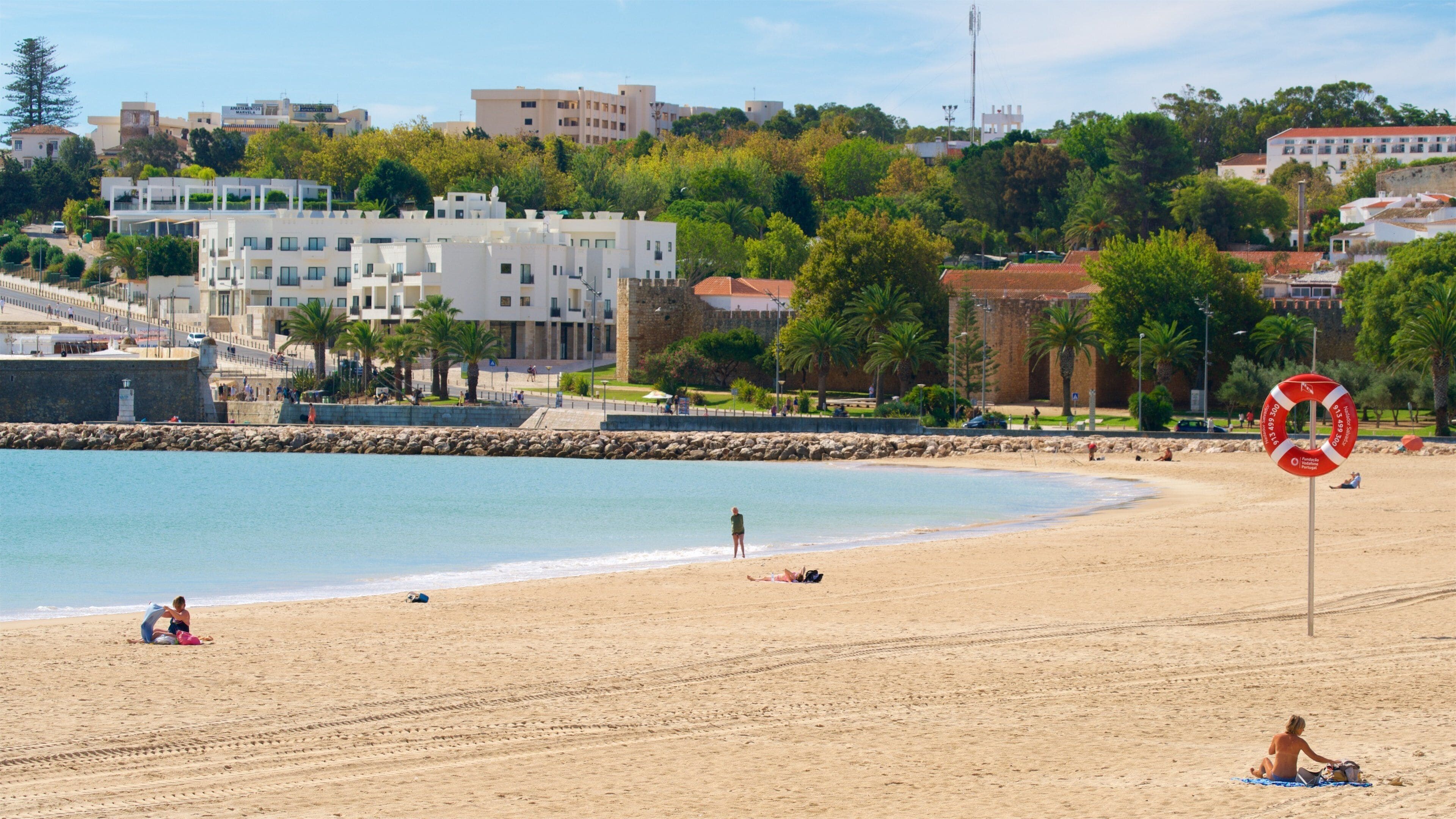 Una playa arenosa con un flotador salvavidas, un muro de piedra y edificios al fondo.