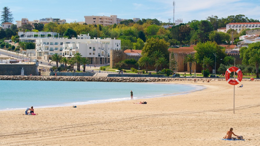 Una playa arenosa con un flotador salvavidas, un muro de piedra y edificios al fondo.