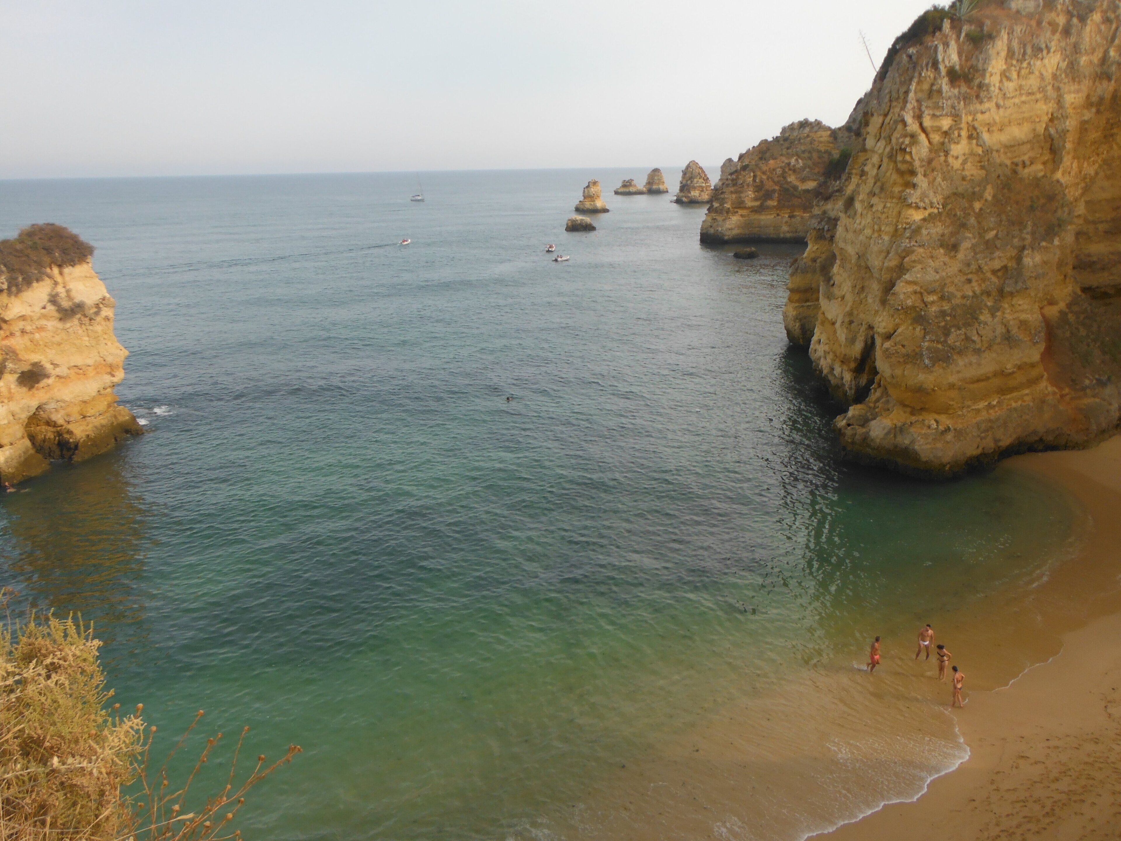 Paisagem costeira com águas claras, falésias rochosas e uma praia arenosa com pessoas.