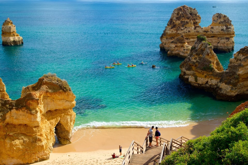 Paisagem costeira com águas turquesa cristalinas, formações rochosas e uma praia de areia com uma passarela de madeira.