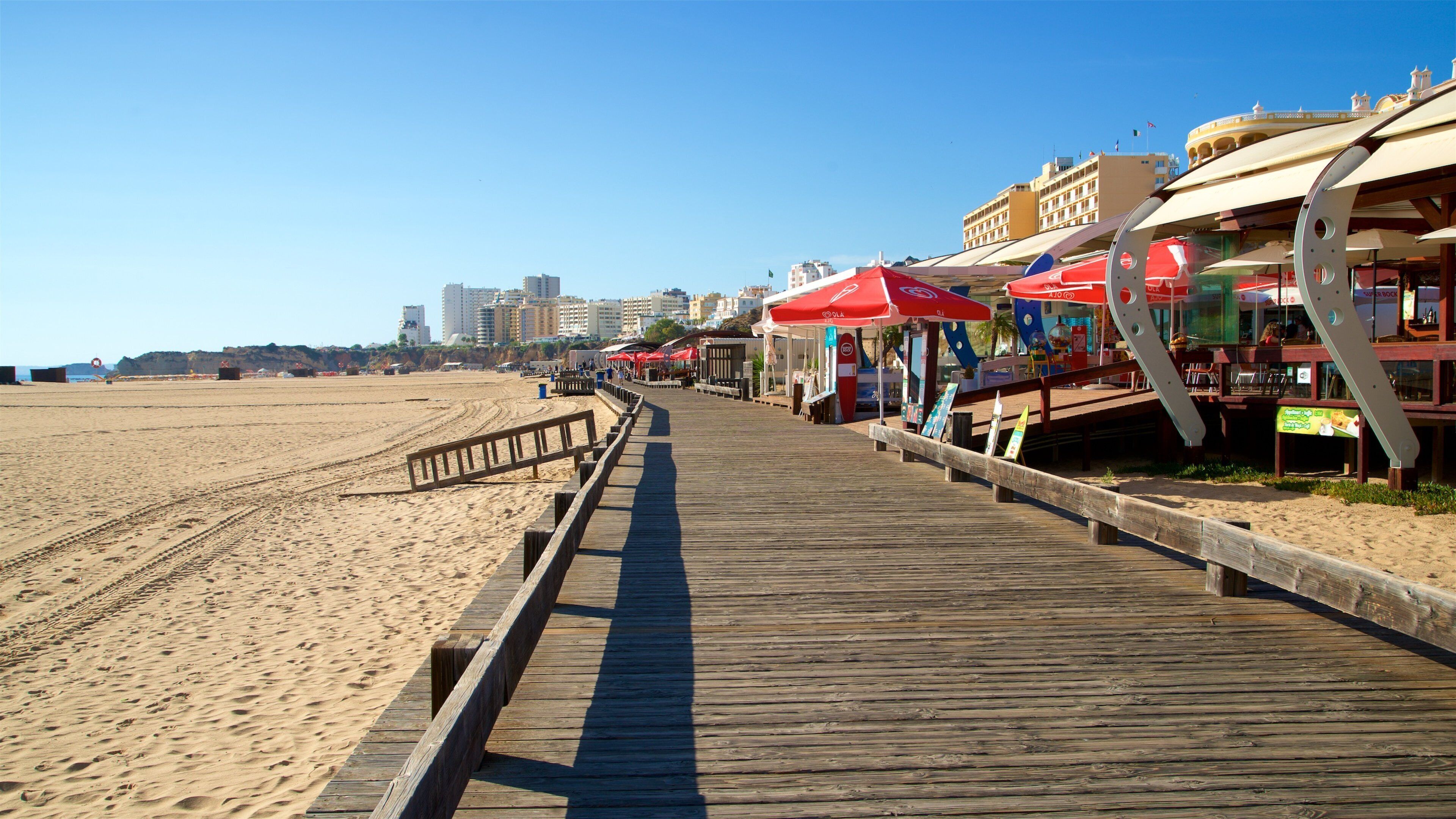 A wooden boardwalk along a sandy beach with red umbrellas and beachside restaurants.