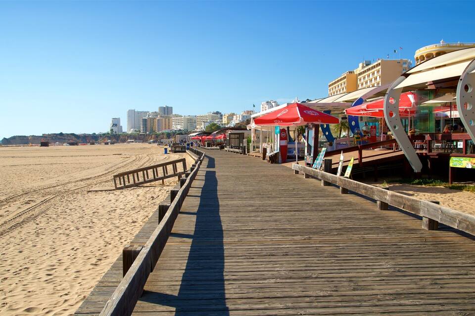 A wooden boardwalk along a sandy beach with red umbrellas and beachside restaurants.