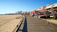 Un paseo marítimo de madera a lo largo de una playa arenosa, con sombrillas rojas y restaurantes frente al mar.