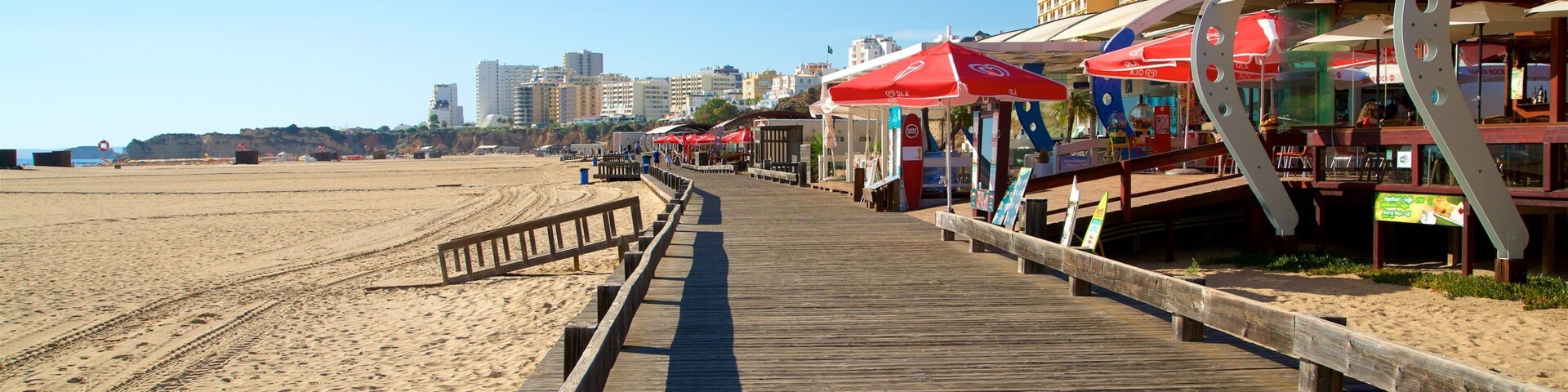 Um calçadão de madeira margeia uma praia arenosa, com guarda-sóis vermelhos e restaurantes à beira-mar.
