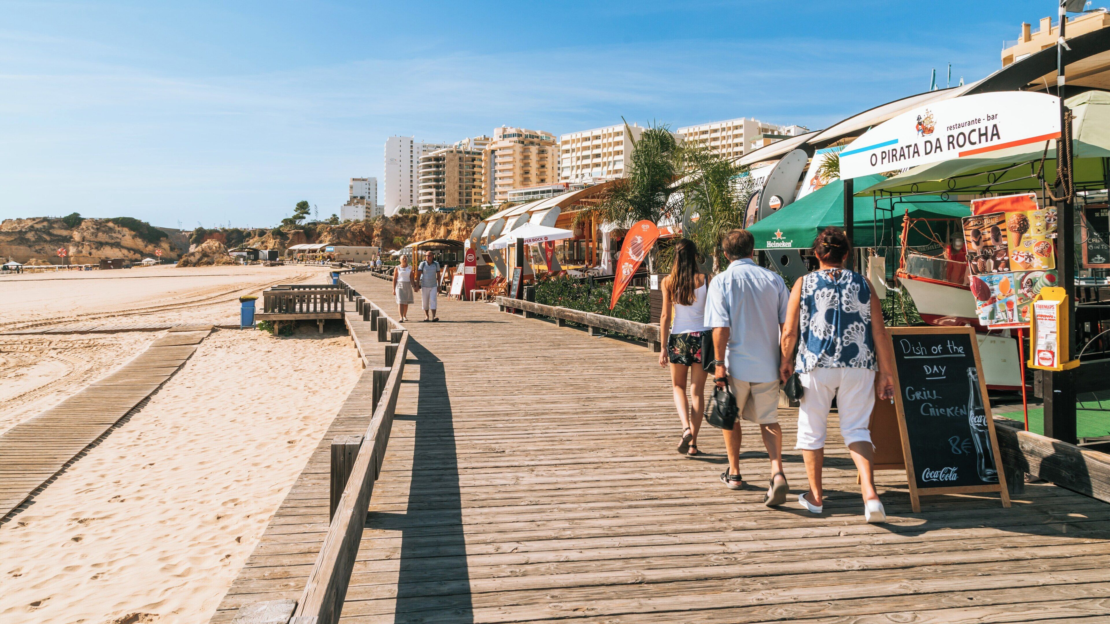Strolling along the boardwalk at Rocha Beach in Praia da Rocha, Faro District, Portugal on a sunny day with colorful cafes and beachgoers