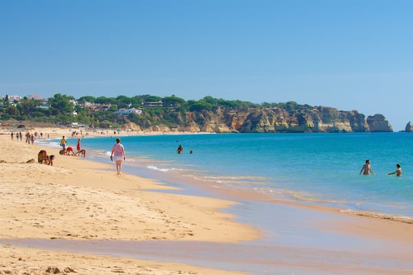 Ein sandiger Strand mit klarem, blauem Wasser, Personen beim Schwimmen und Spazierengehen, und felsige Klippen im Hintergrund.