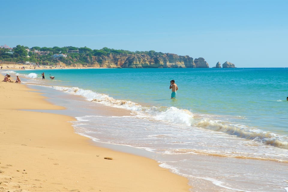 A sandy beach with gentle waves, a clear blue sky, and distant rocky cliffs.