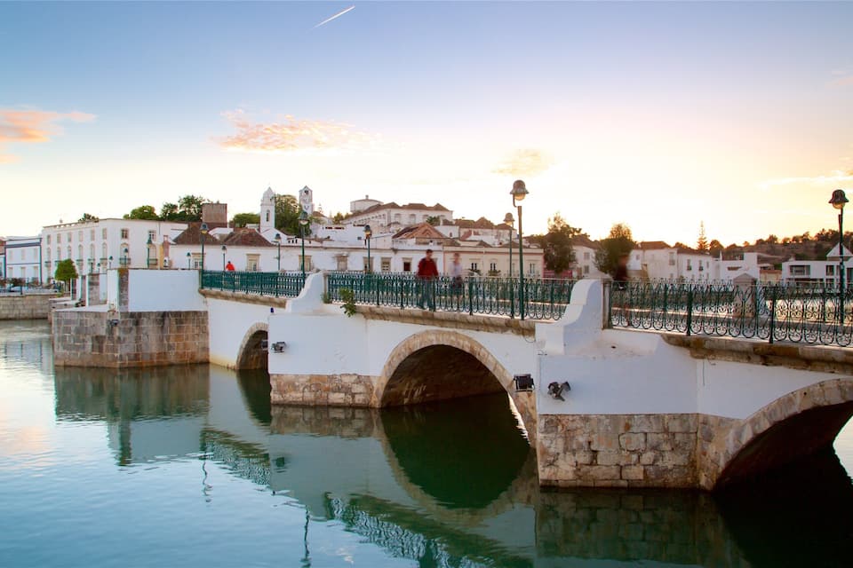 A stone bridge over a calm river, with buildings and a sunset in the background.