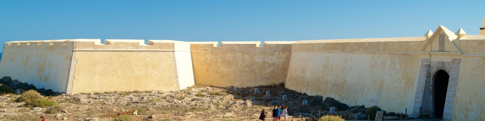 Uma grande fortaleza com um arco central, rodeada por terreno rochoso e vegetação escassa.