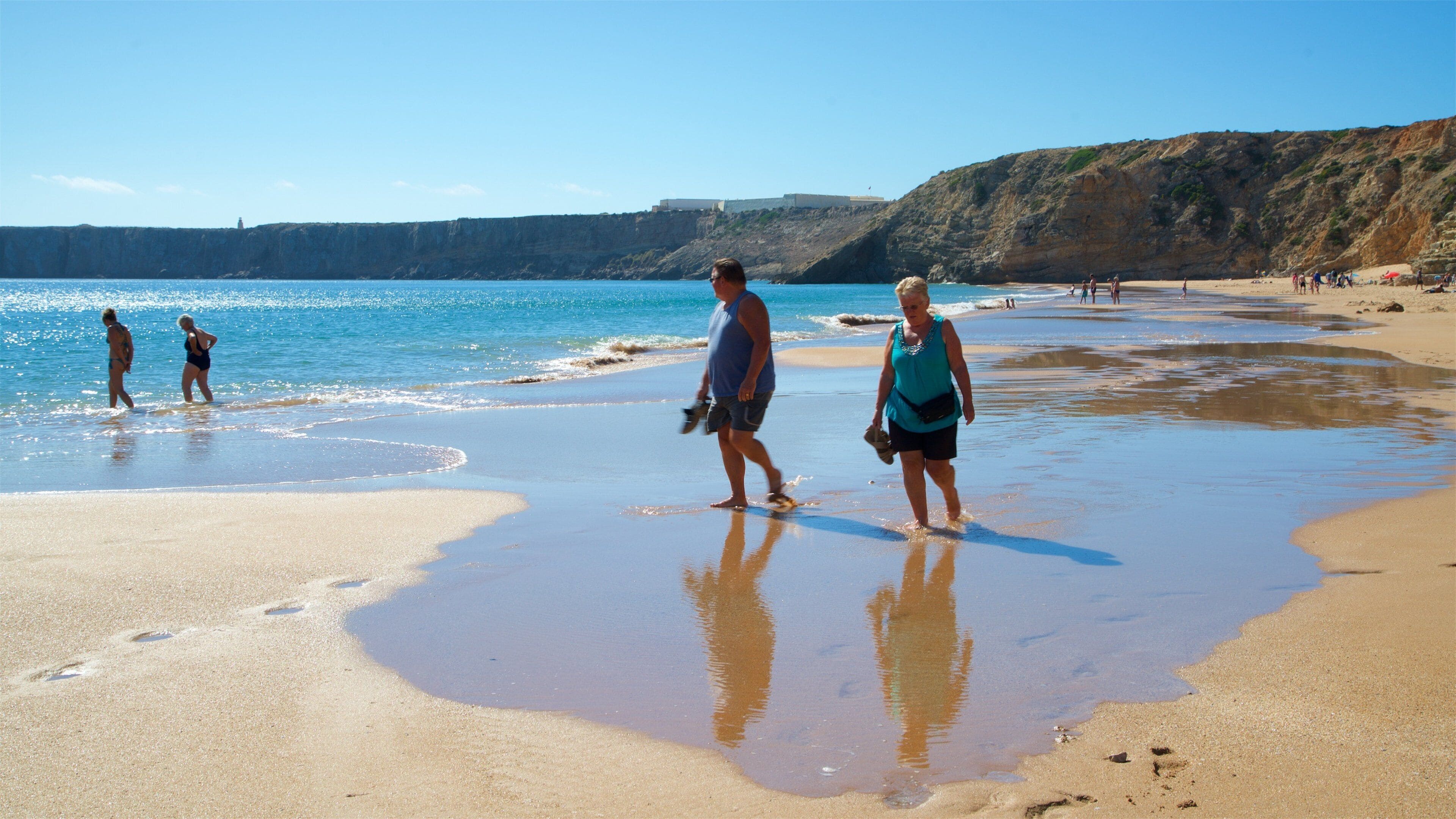 A beach with clear blue water, sandy shore, and rocky cliffs in the background.