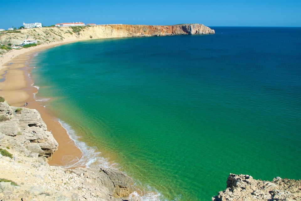 A coastal landscape with a clear blue sea, a sandy beach, rocky cliffs, and a few buildings on a hillside.