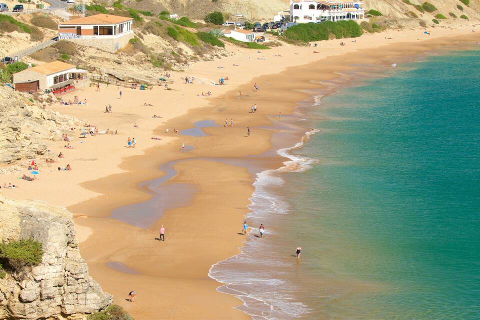 A sandy beach with clear turquoise water, rocky cliffs, and a few buildings in the background.