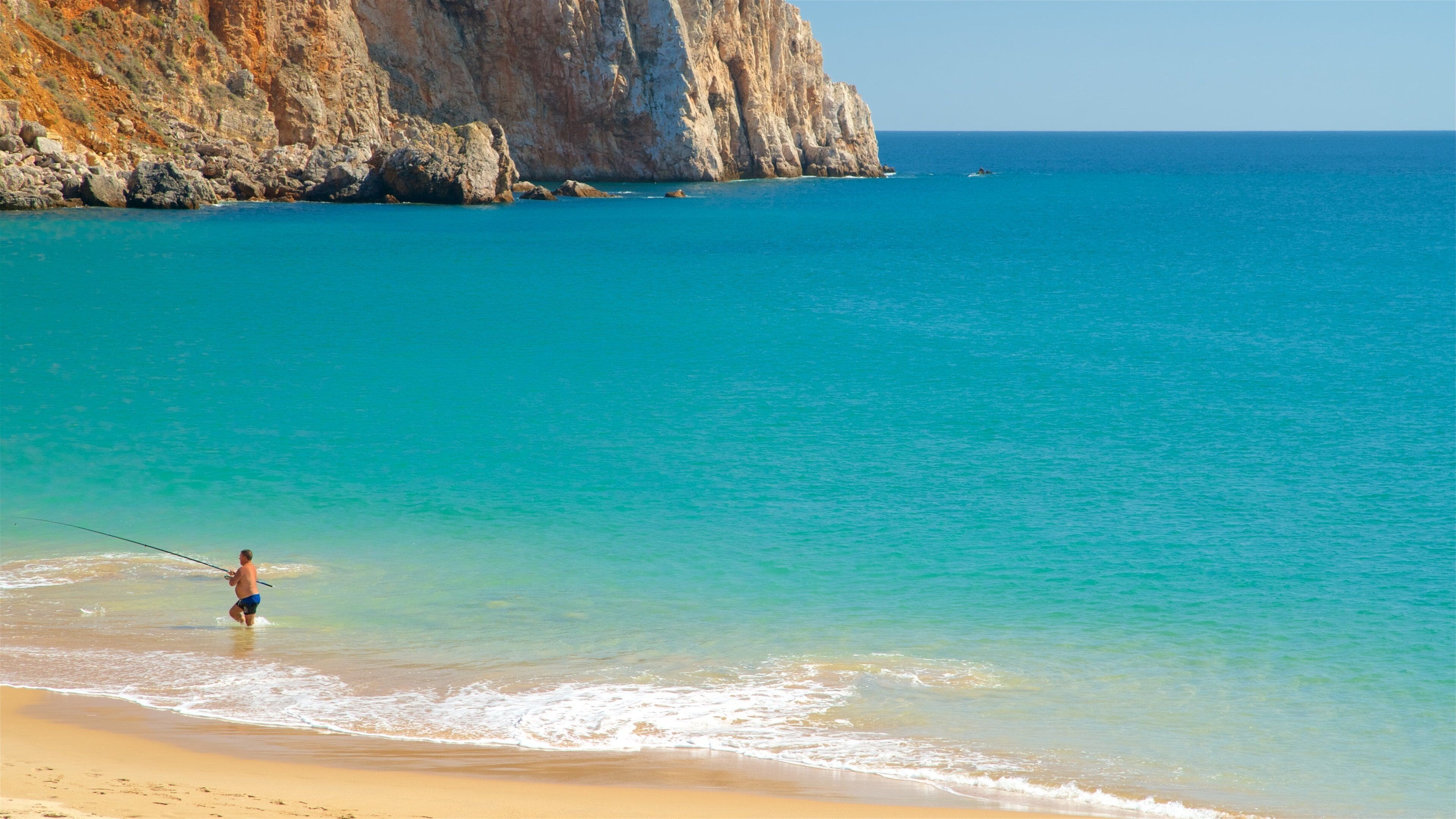 A beach with clear turquoise water, a person fishing, and rocky cliffs in the background.