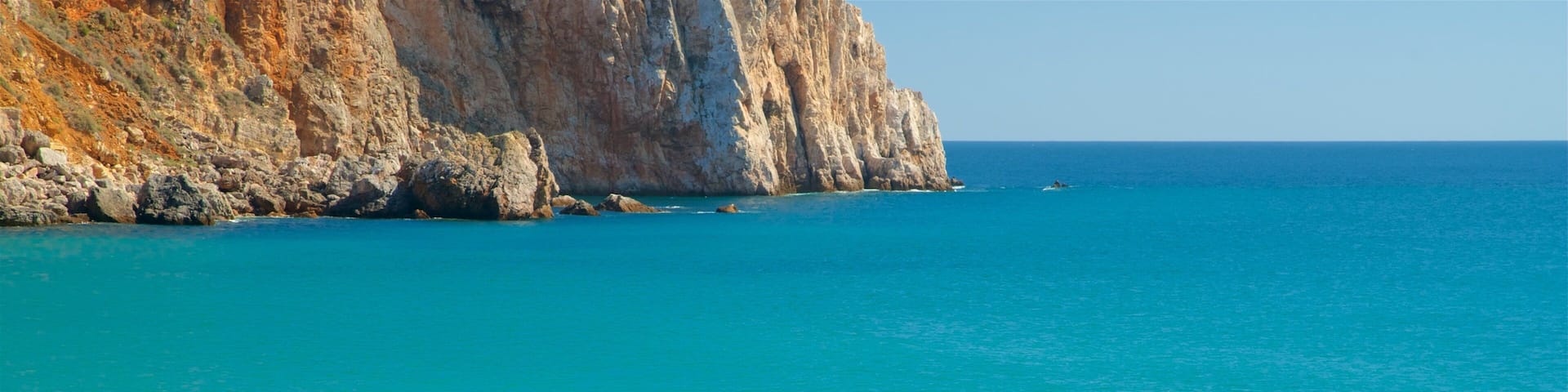 A beach with clear turquoise water, a person fishing, and rocky cliffs in the background.