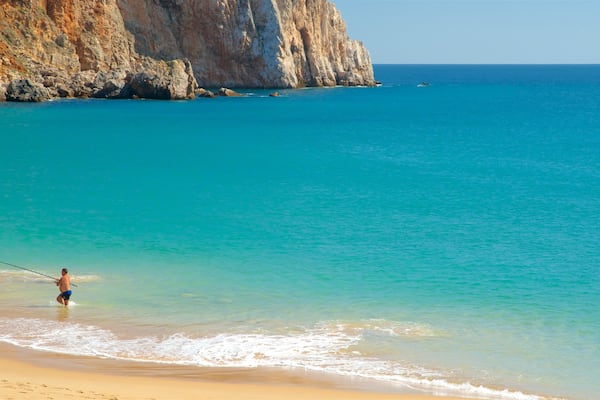 A beach with clear turquoise water, a person fishing, and rocky cliffs in the background.
